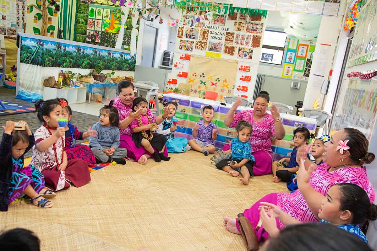 A group of children and kaiako sitting around the whāriki singing and playing instruments.