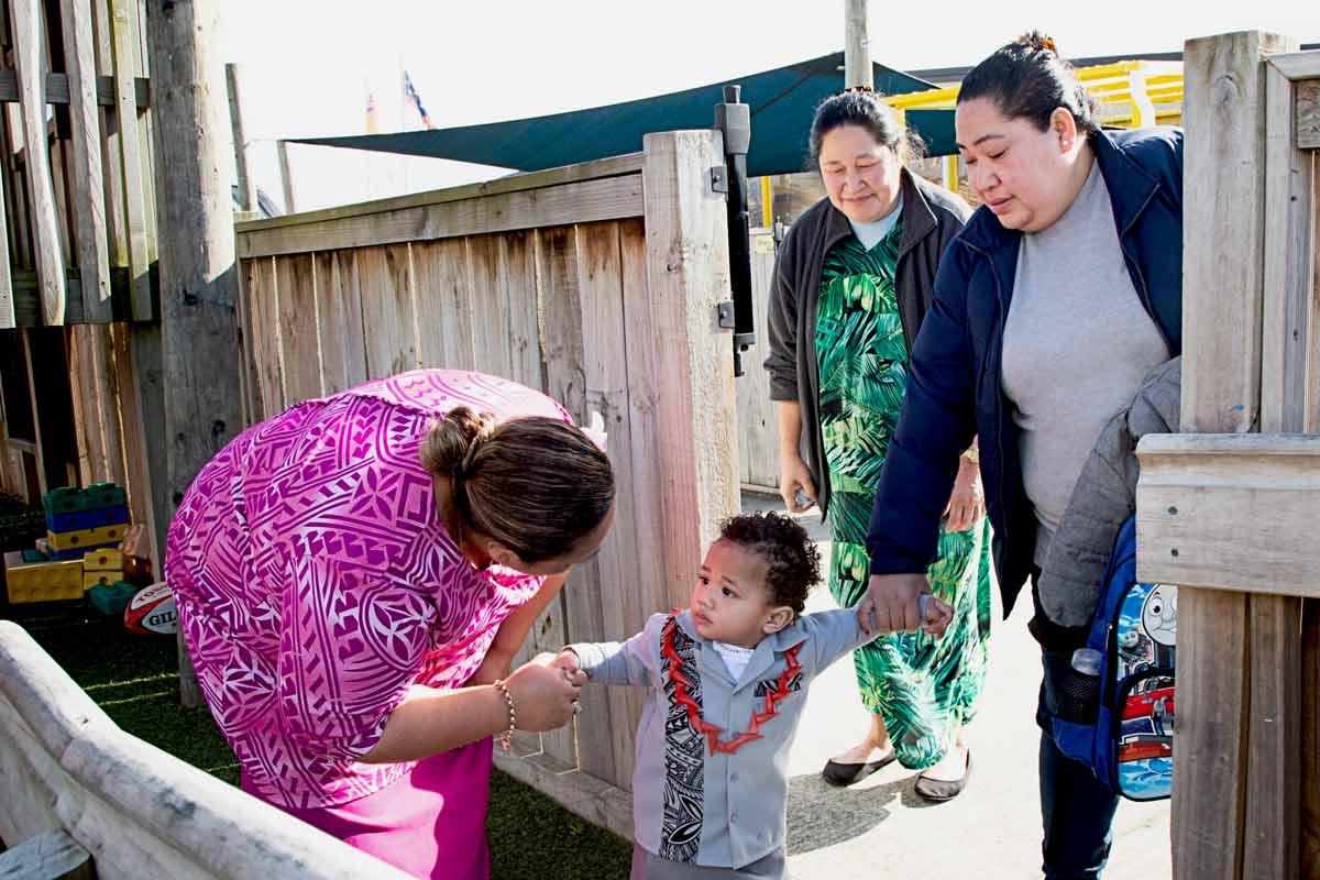 Child being dropped off at preschool and the teacher meeting the child and the parents at the gate.