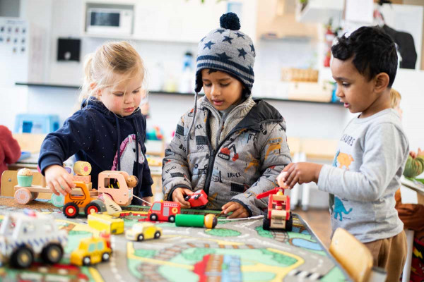 Tamariki play together with vehicles on a mat, a person shaped toy lies down. 