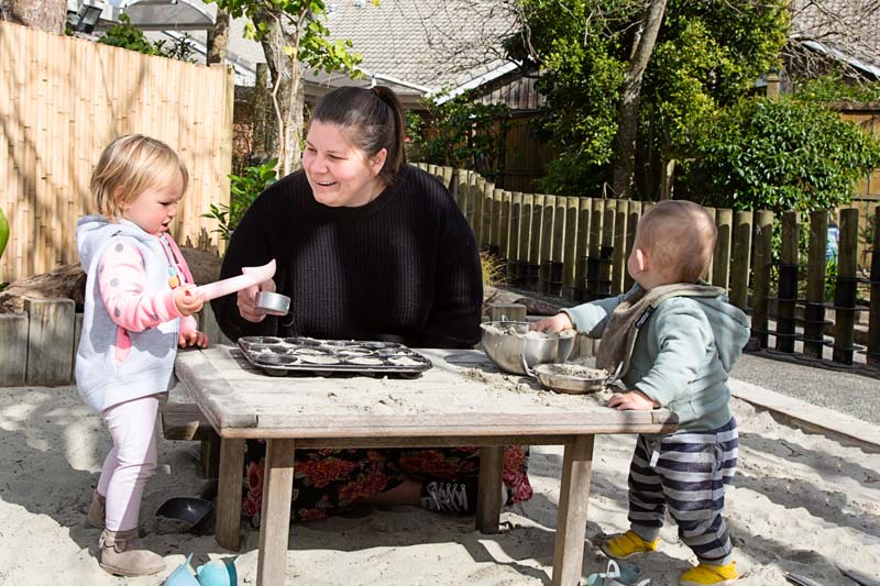 A kaiako talks with two tamariki while playing with bowls and sand.